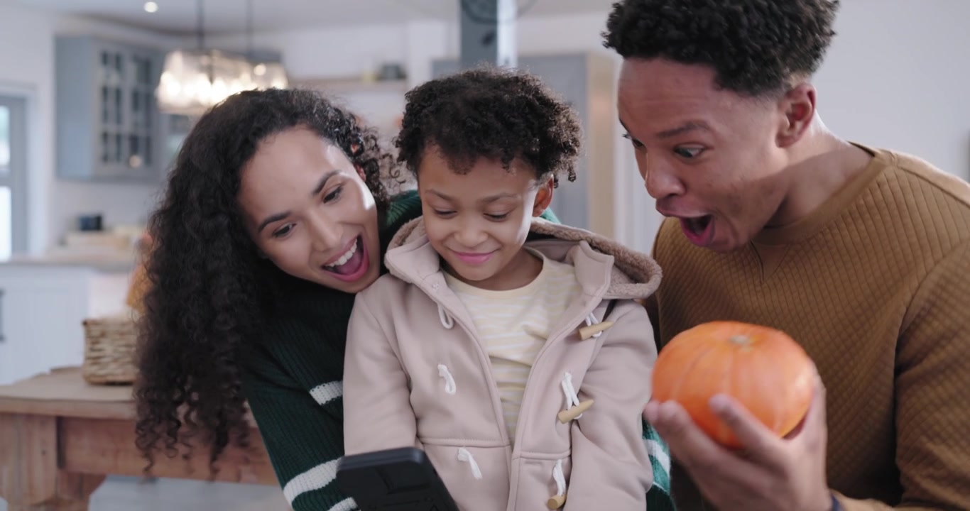 Cucina di famiglia afroamericana, mamma che punta il telefono nero per un selfie, papà che saluta con una zucca.