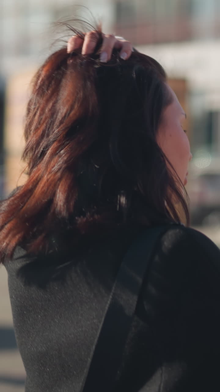 드폰을 들고 밖에서 걸어다니는 여성이 머리를 조정하고, 장치에 다시 초점을 맞추기 전에 잠시 주위를 둘러보고, 배경에는 현대적인 도시 건축물과 녹색 자전거가 있습니다.