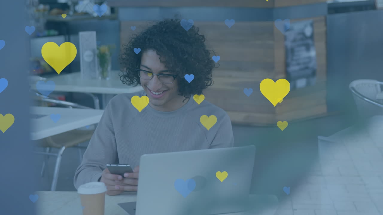 animación de iconos de corazón amarillo y azul sobre un hombre biracial usando un teléfono inteligente en una cafetería