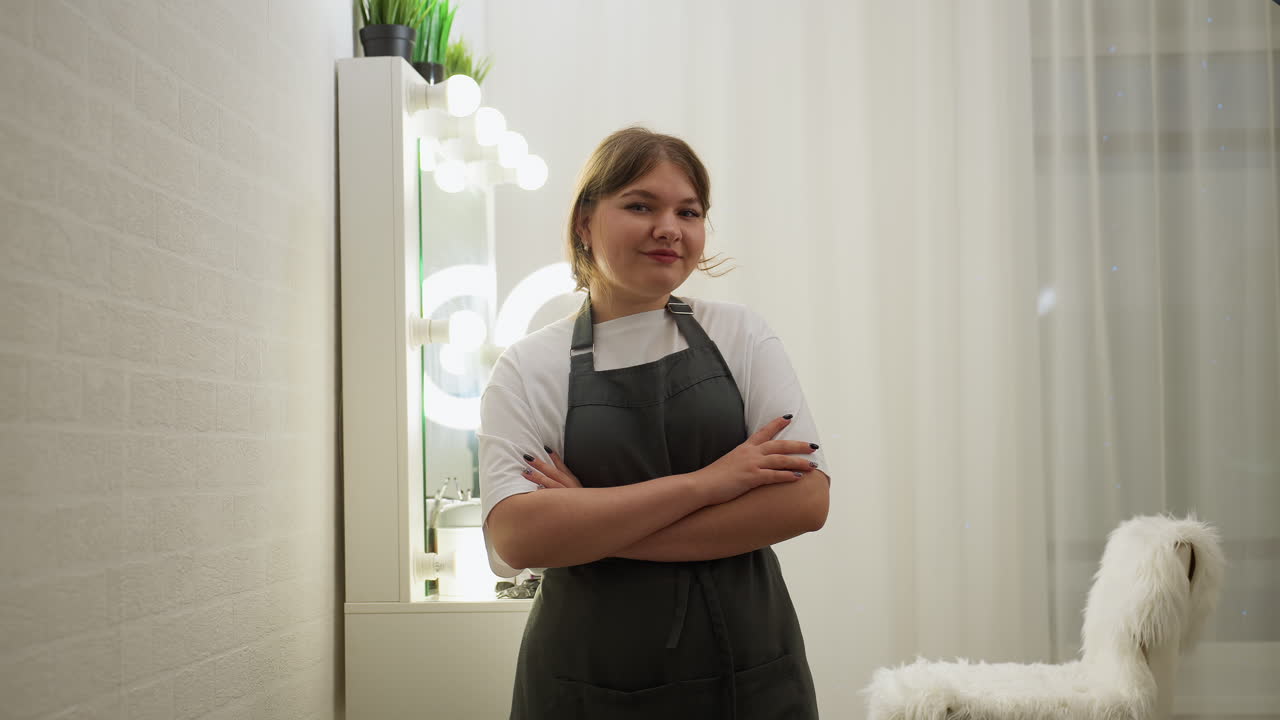 Young woman with short blond hair stands confidently with folded arms, smiling softly in stylish salon interior featuring soft lighting, modern vanity mirror with bulbs, and white furry chair
