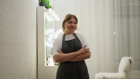 young woman with short blond hair stands confidently with folded arms, smiling softly in stylish salon interior featuring soft lighting, modern vanity mirror with bulbs, and white furry chair