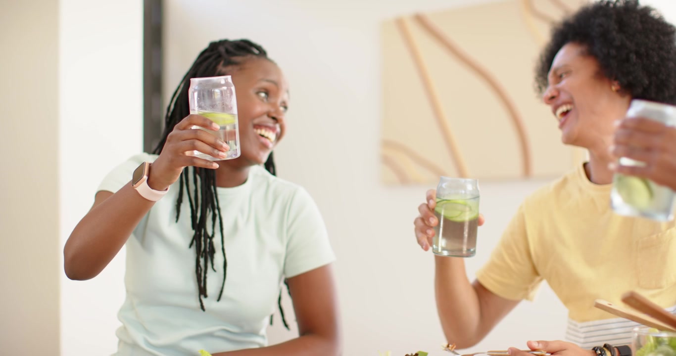 Femmes afro-américaines trinquant avec des verres contenant du concombre à table, encourageant une troisième femme à les rejoindre, sirotant.
