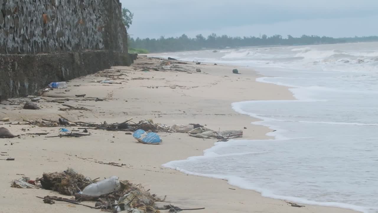 Olas que llegan a una playa de arena cubierta de basura, residuos plásticos y escombros, en cámara lenta y toma amplia