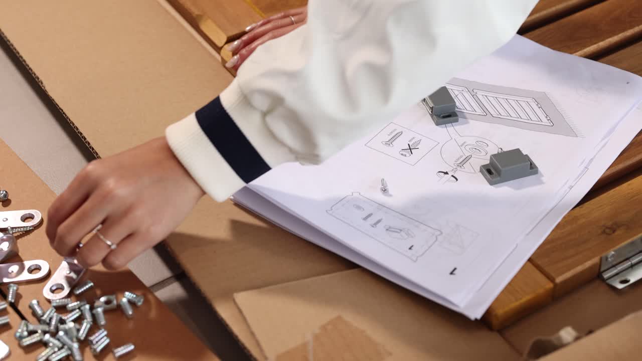 Hands organize parts and follow instructions for furniture assembly on a wooden surface with natural lighting