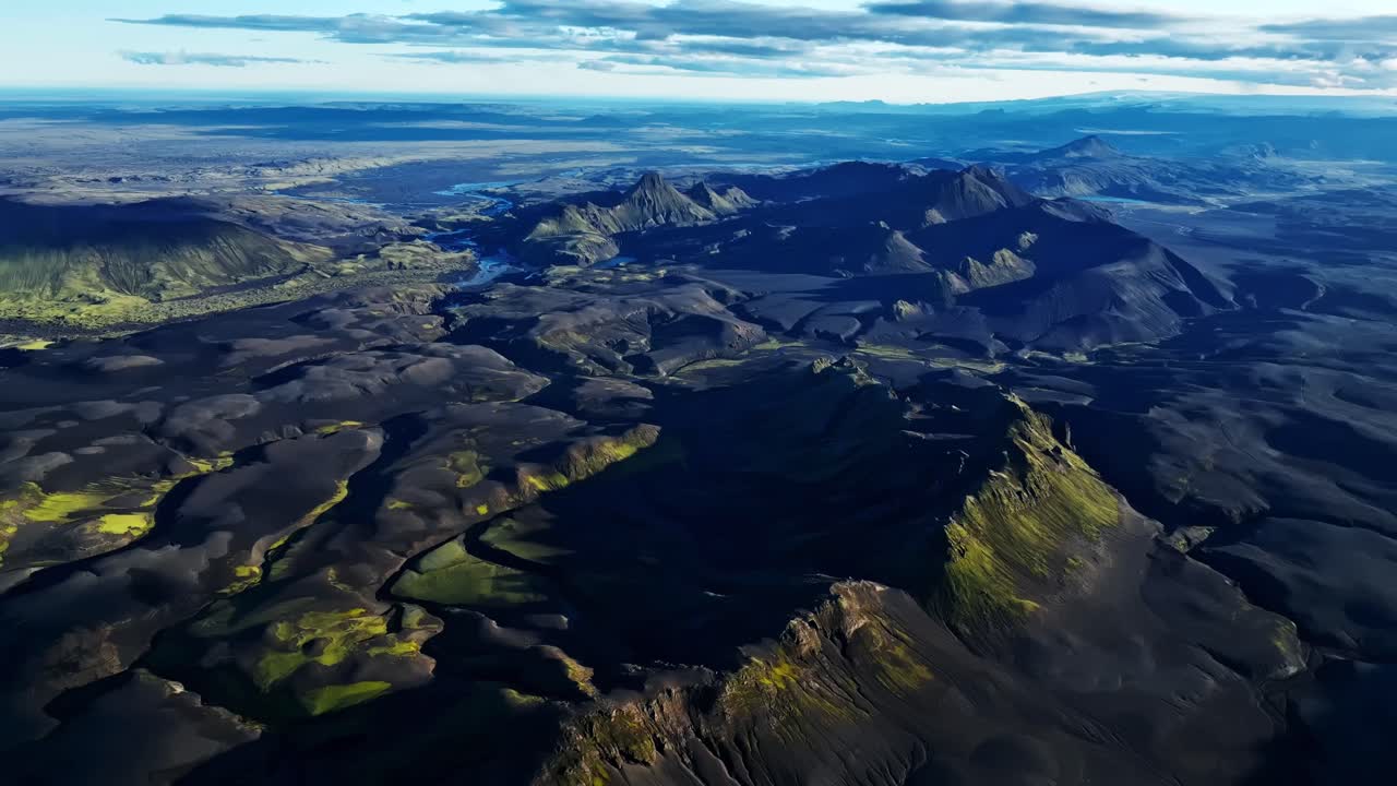 Wide aerial view of Iceland’s volcanic highlands, showing sprawling black lava plains, fractured ridges and scattered moss textures stretching across a raw, ancient eruption landscape