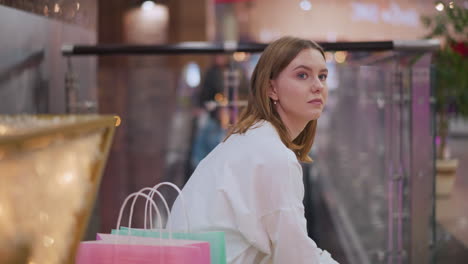 lady seated on bench in shopping mall holding tablet surrounded by festive decorations, illuminated star decor, shopping bags beside her, with blurred figure on moving escalator in vibrant