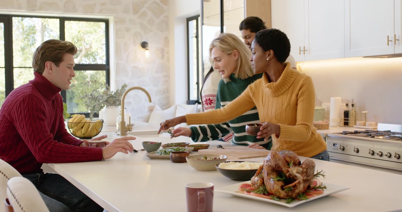 Diverse group mustard-knit woman serving and passing turkey bowls at kitchen island preparing meal