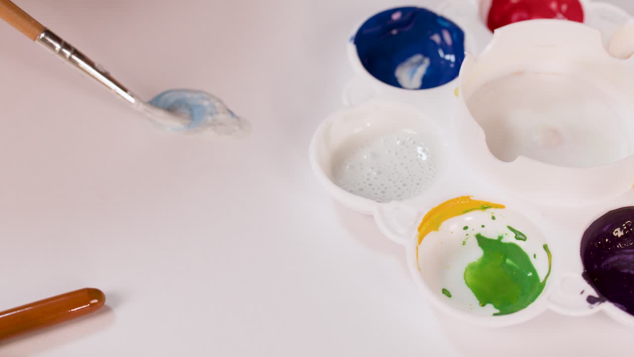 Hand mixes blue watercolor paint with brush on palette, soft lighting, overhead close-up view