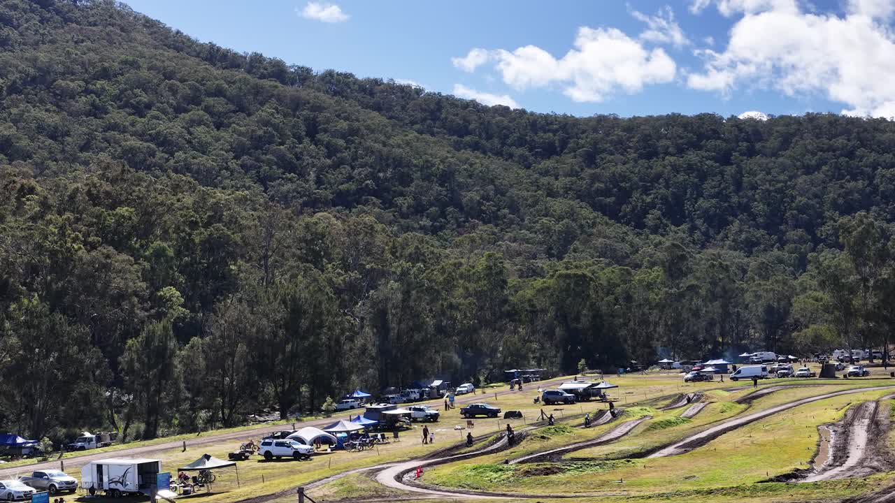 Motocross riders traverse winding dirt track under bright daylight, surrounded by forested hills and vehicles