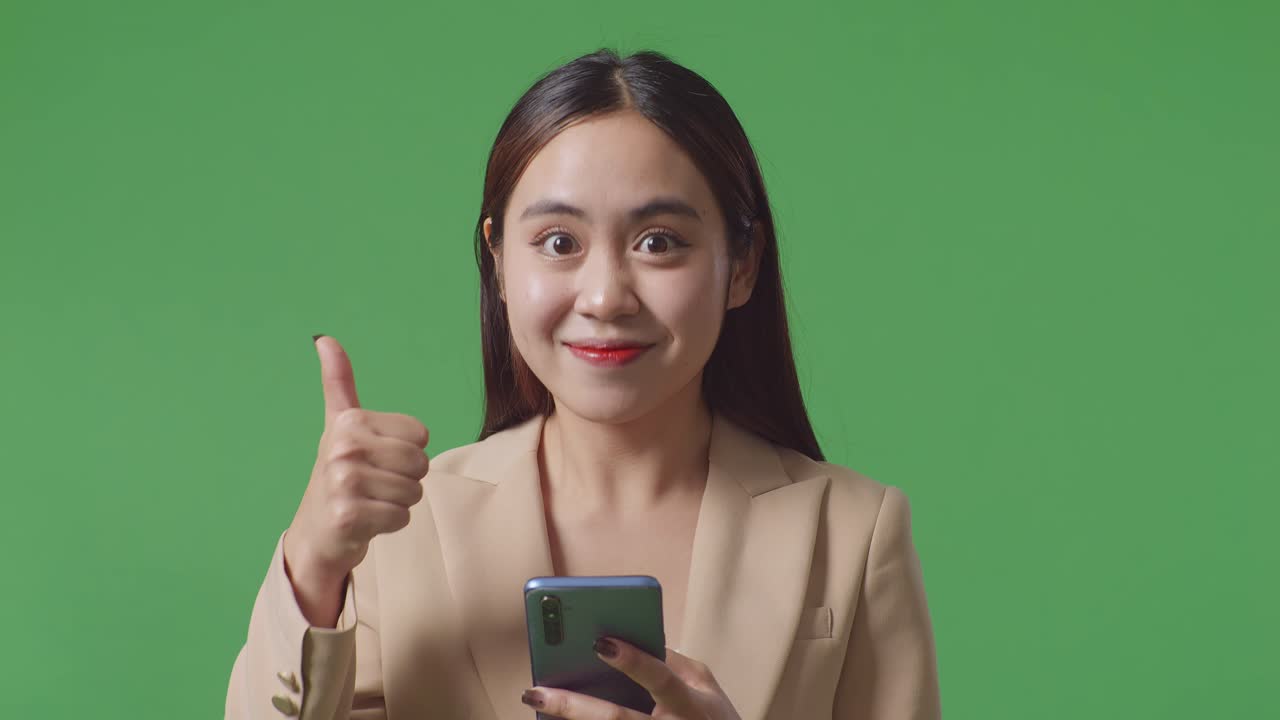 스마트폰을 사용하는 아시아 사업가 여성의 근접 사진과 스튜디오에서 녹색 화면에 서있는 동안 엄지손가락을 보여주는