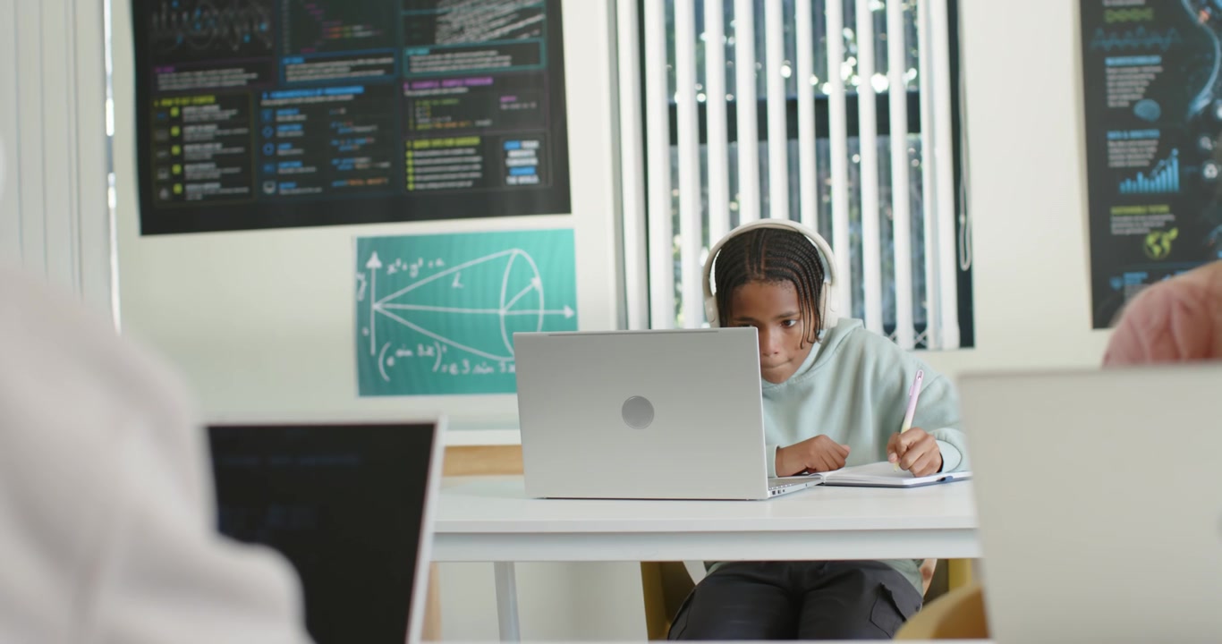 African American teen student in class using laptop with headphones taking notes lecture starting