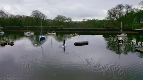 Beautiful-aerial-view,-footage-of-middle-aged-man-paddle-boarding-on-Rudyard-Lake-in-the-Derbyshire-Peak-District-National-Park,-popular-holiday,-tourist-location-with-peaceful-calm-water