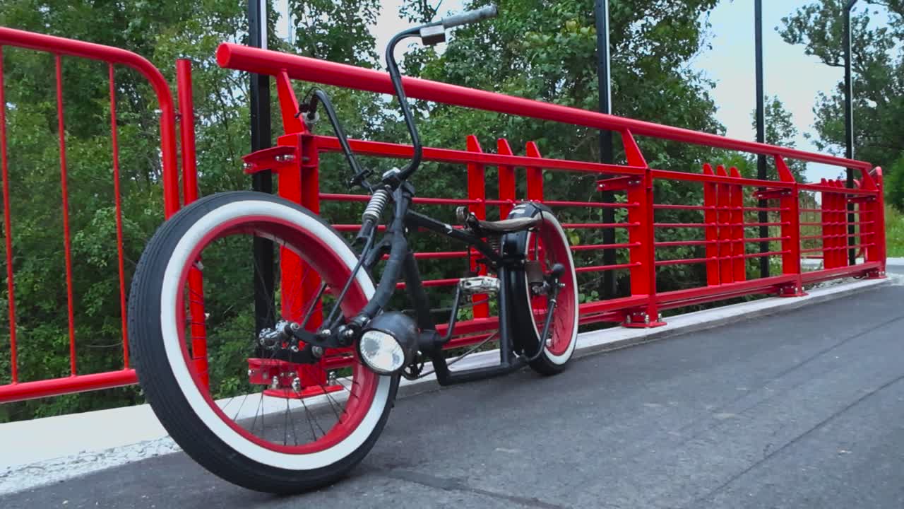 Footage of a black chopper styled bike or a bicycle with whitewall wheels and red rims on a bridge with red railings