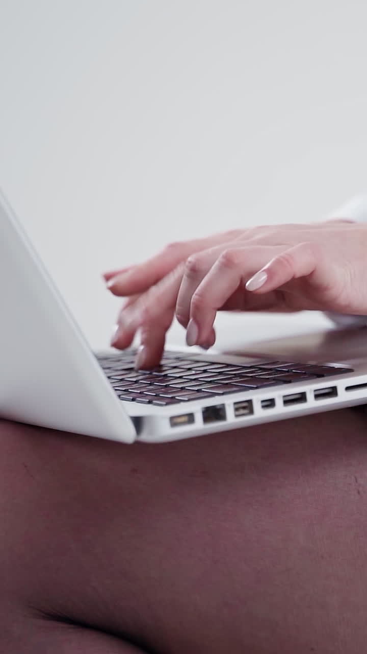 Young female is typing on a laptop. White laptop on woman's knees. Hands of a woman working on a modern gadget. Close-up. Slow motion. Vertical video