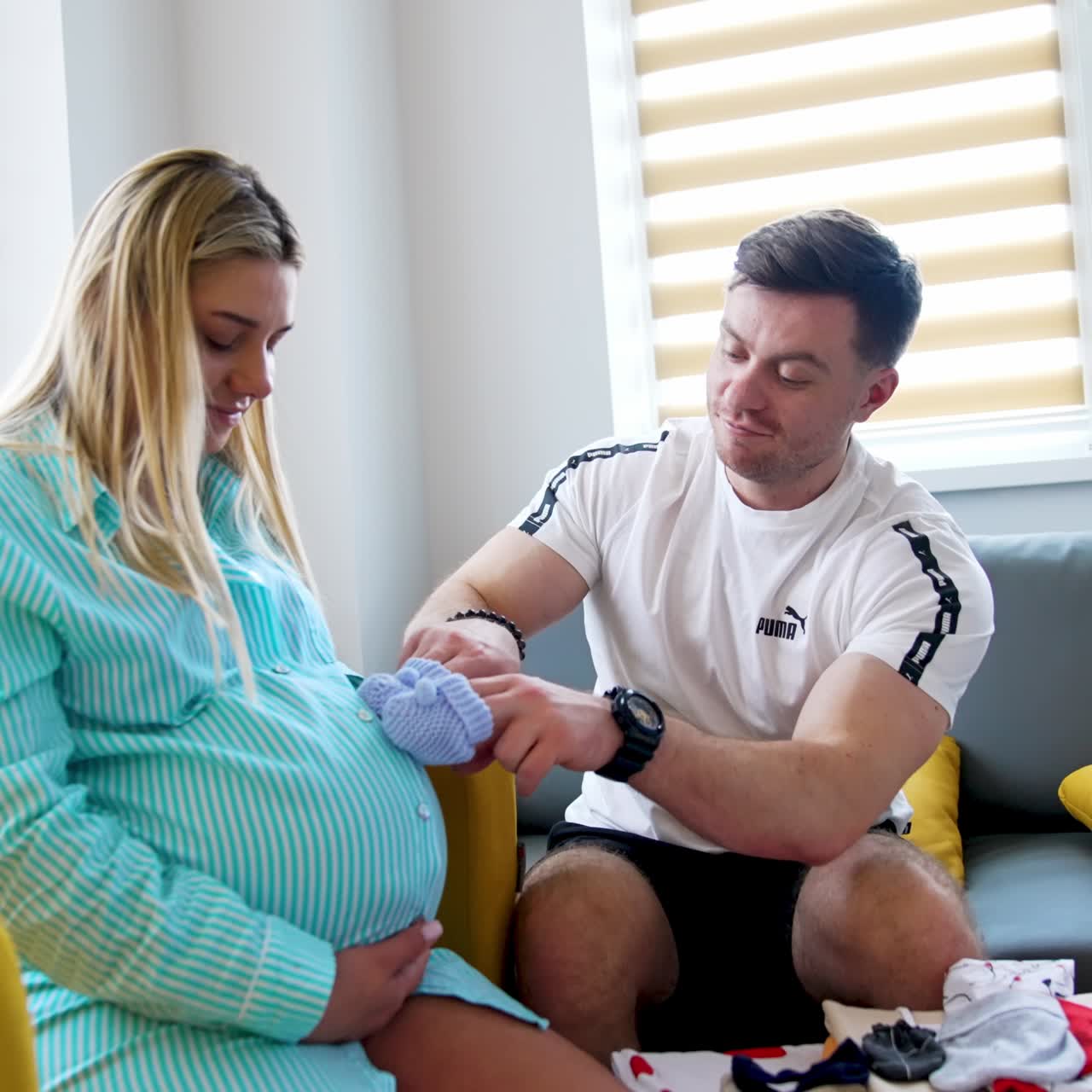 Blonde woman with big belly sits on the sofa. Her husband takes little baby socks and steps them gently by the abdomen. Happy expecting couple in the hospital
