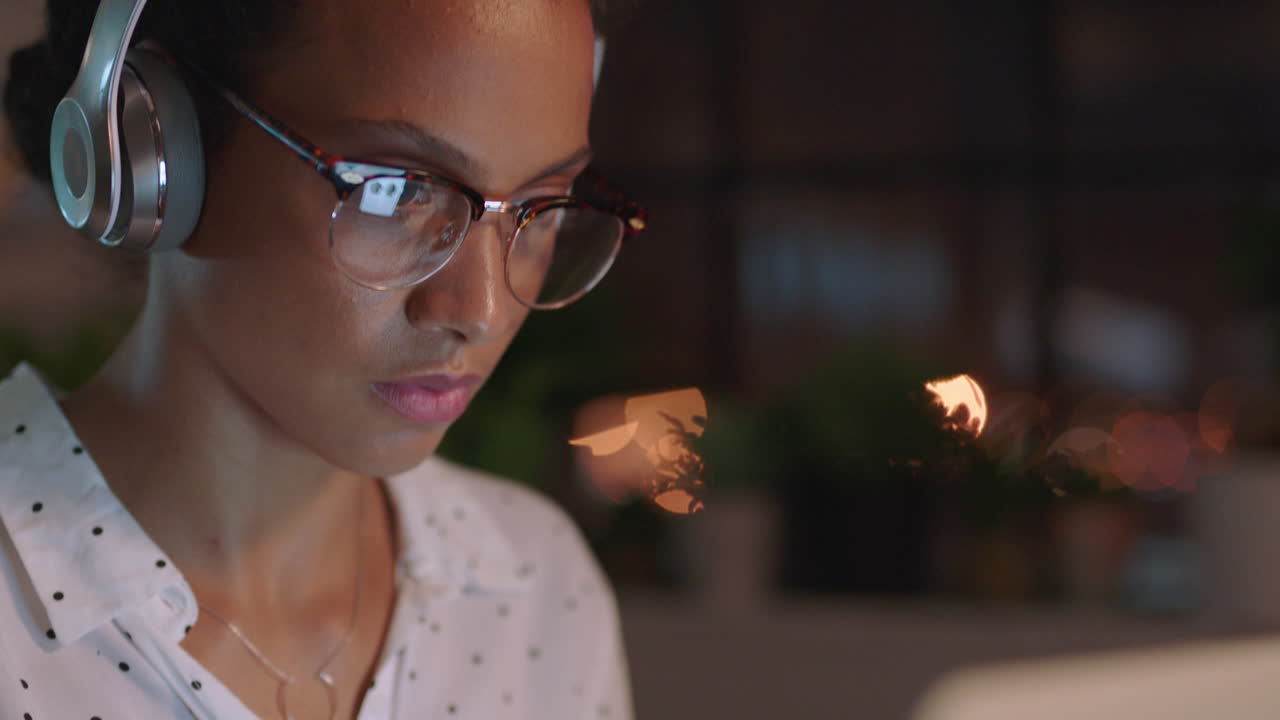 mujer de negocios de raza mixta que trabaja hasta tarde usando una computadora portátil escribiendo gerente de proyecto navegando en línea planeando la investigación fecha límite escuchando música en la oficina por la noche