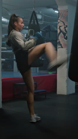 female kickboxer in boxing bandages hits punching bag while training in dark boxing gym. athletic woman practices and prepares for match with male coach. vertical shot