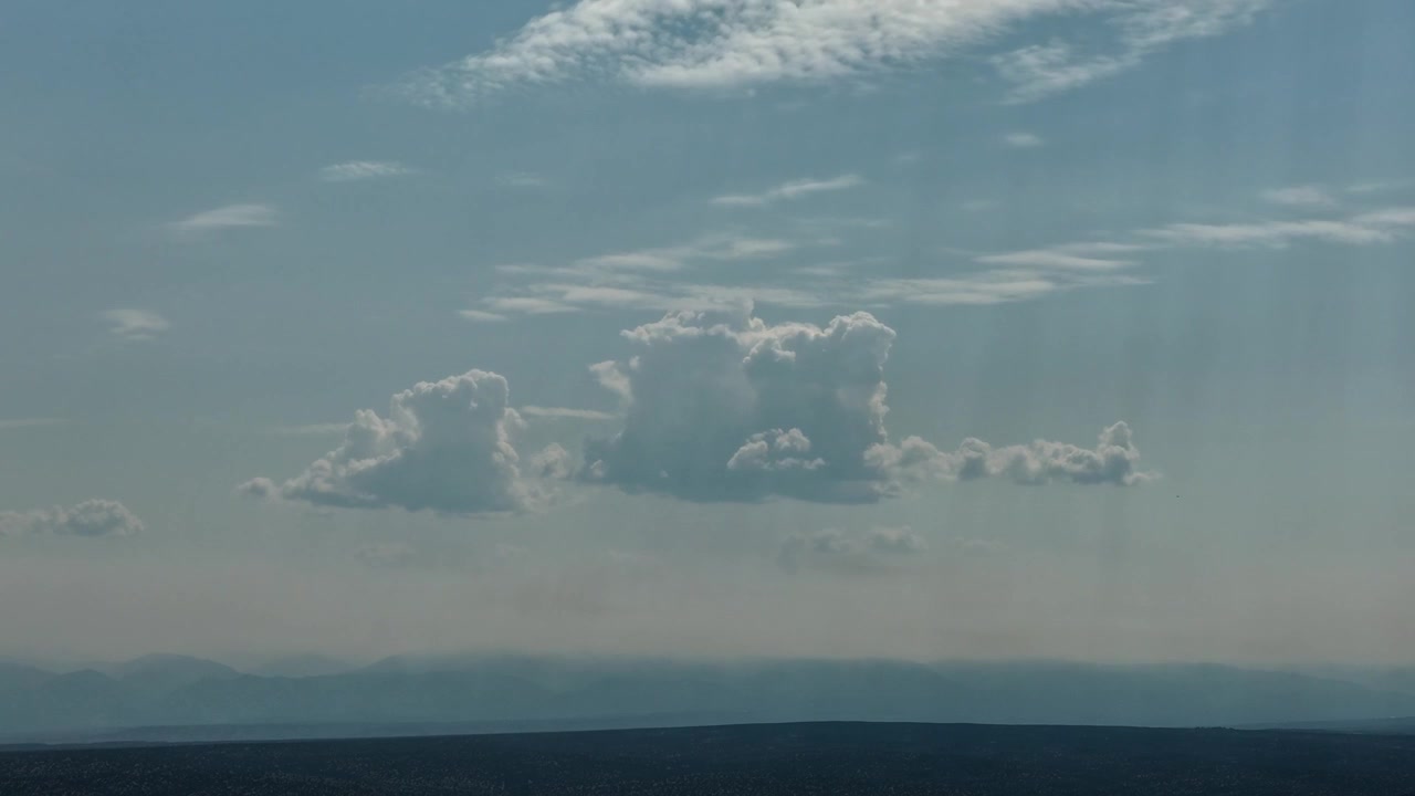 4K aerial timelapse of cumulus clouds shifting in place above a dark mountain range. A thick layer of wildfire smoke settles over the landscape, casting a moody blue haze across the scene.