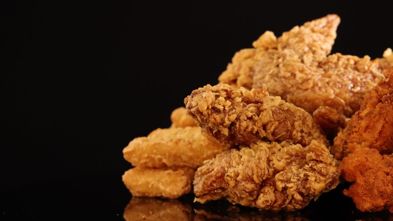 Golden fried chicken wings, drumsticks, and nuggets rotate on a reflective black surface under warm, focused lighting, highlighting crunchy textures and appetizing detail