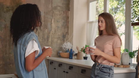 black girl and caucasian young woman talk and laugh. two female friends have a cup of coffee in the kitchen. medium shot.