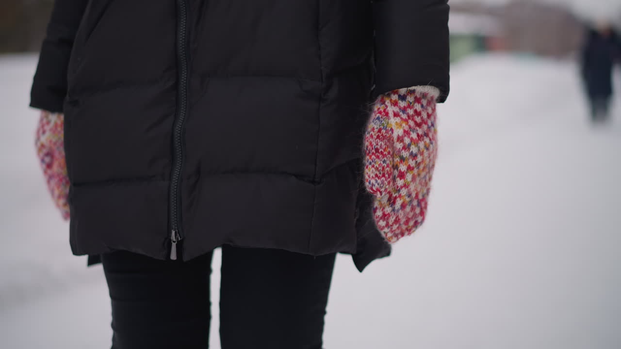Female wearing black winter coat walking outdoors in snowy environment with colorful knitted mitten visible close up hand detail cold season background blurred people distant frosty urban lifestyle scene