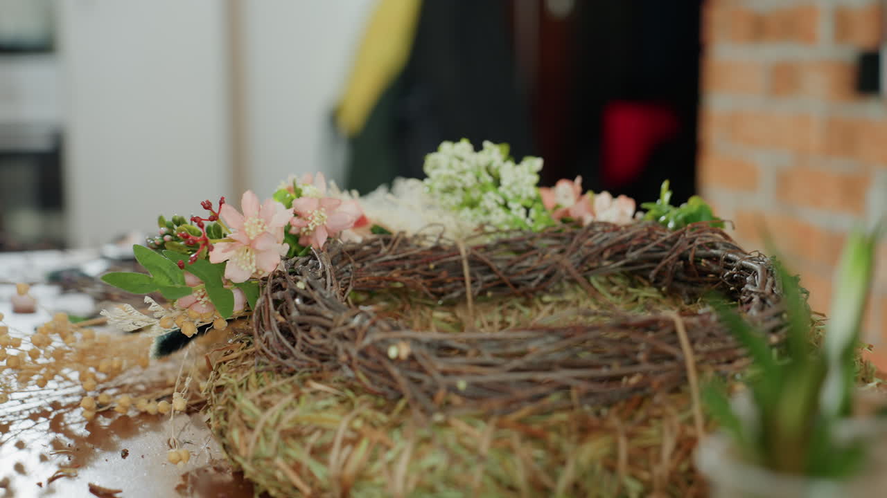 Close up of handmade wreath decorated with pink flowers, green leaves placed on wooden table surrounded by twine, moss, and plants, highlighting rustic floral craftwork and artistic decoration process