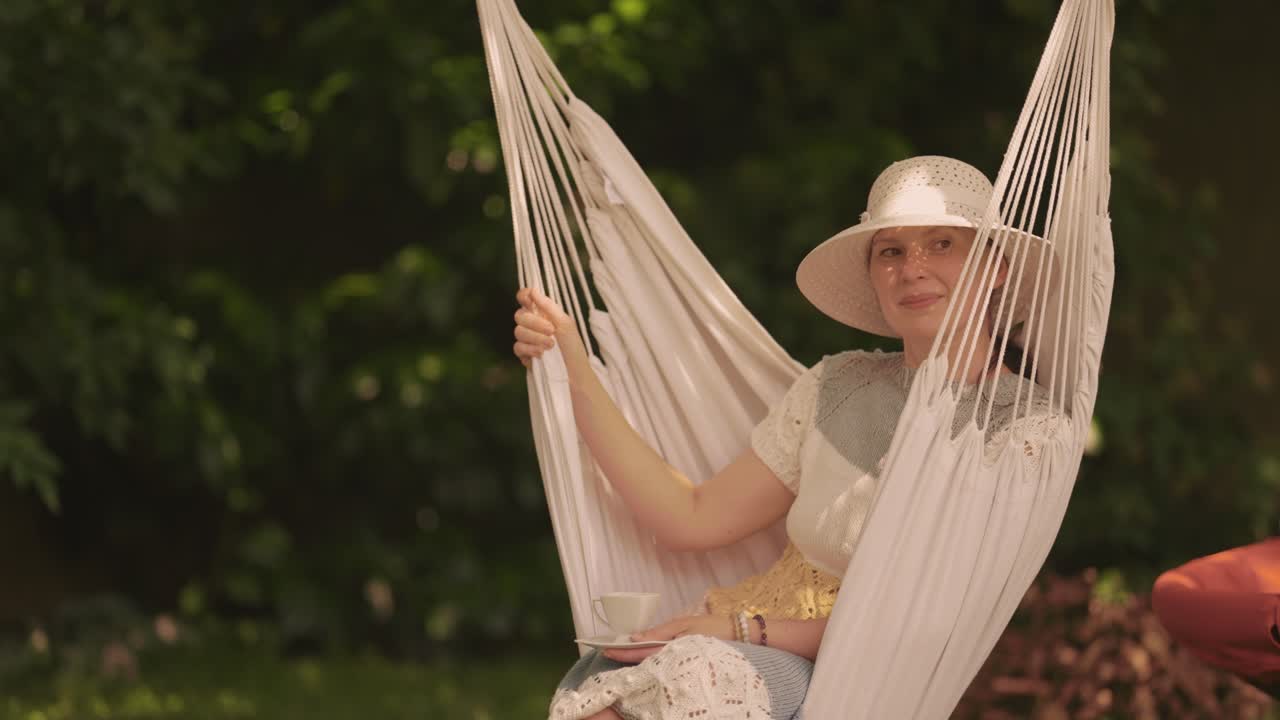una mujer de mediana edad de aspecto casual, con un vestido de punto y un sombrero para el sol, sentada en la hamaca