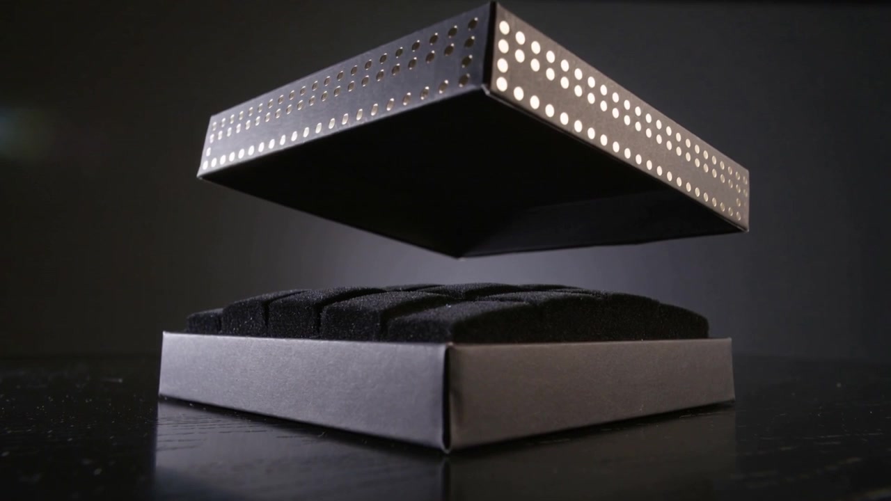 Lifting two-piece black display box, hidden latch releasing lid on glossy tabletop, revealing foam