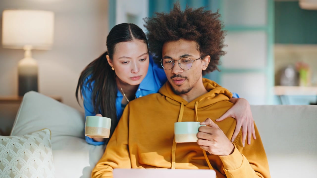 Interracial couple browsing laptop on sofa at home. Woman embracing boyfriend.