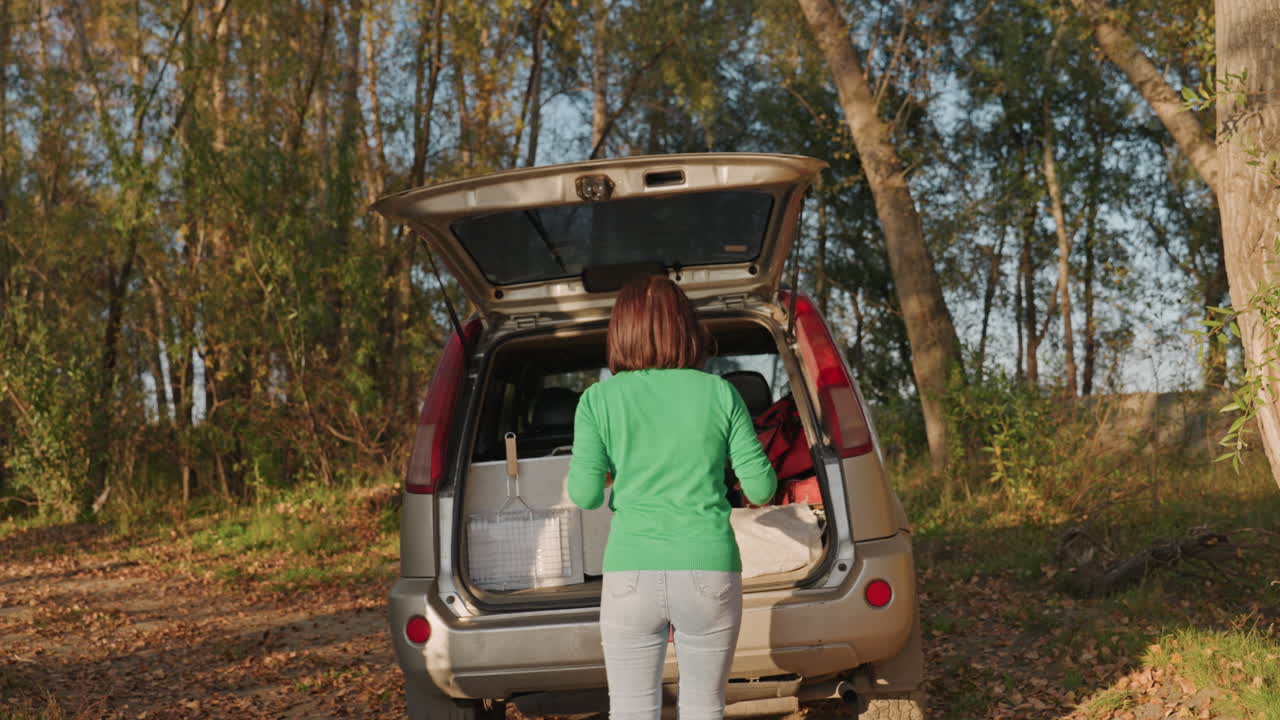 Black woman packing car at campsite opens trunk of suv and loads cooler and boxes, bends and lifts items while organizing gear, surrounded by birch trees and fallen leaves, daylight and brisk autumn