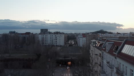 Vista-Aérea-De-Personas-Caminando-Y-Vehículos-Moviéndose-Con-Autos-Estacionados-En-Un-Lado-De-La-Calle-Rodeado-De-Edificios-Y-árboles-Durante-Una-Mañana-Nublada-En-Berlín,-Alemania