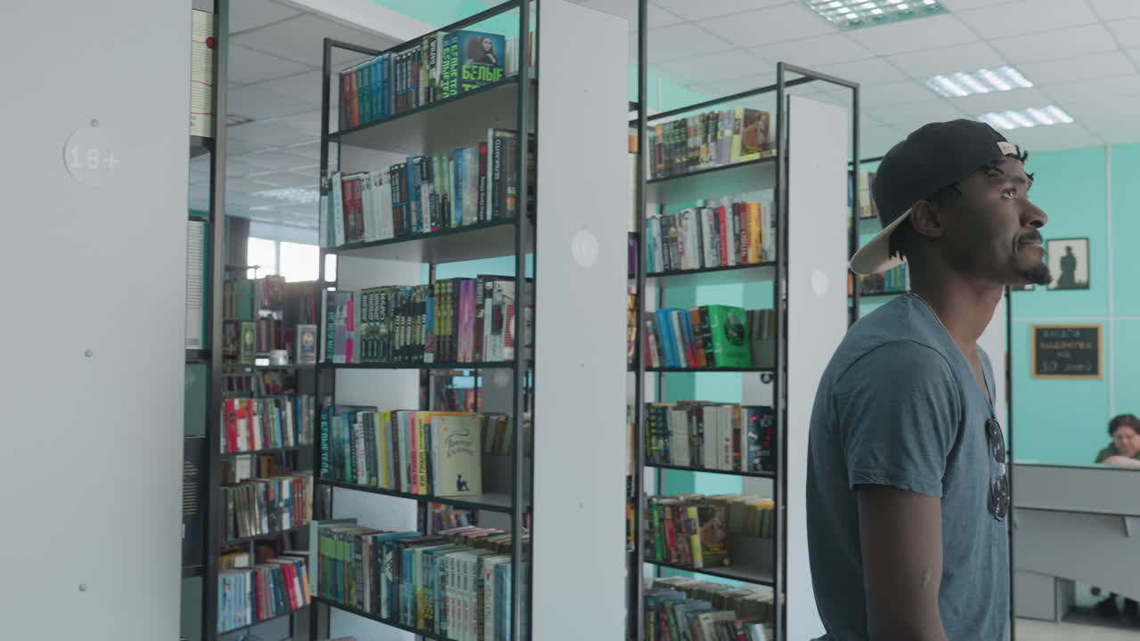 Young man wearing cap and casual shirt standing between library bookshelves, looking around with thoughtful expression while surrounded by rows of colorful books in quiet academic setting