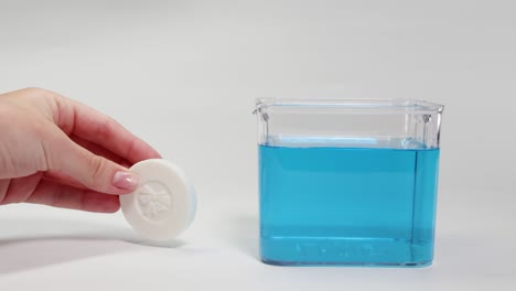 a hand drops a soap bar into a clear container of blue water, observing its sinking behavior