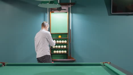 gentleman in white shirt and grey trousers walking into billiard room towards billiard ball rack. green walls, pool table in background, overhead light, classic decor environment