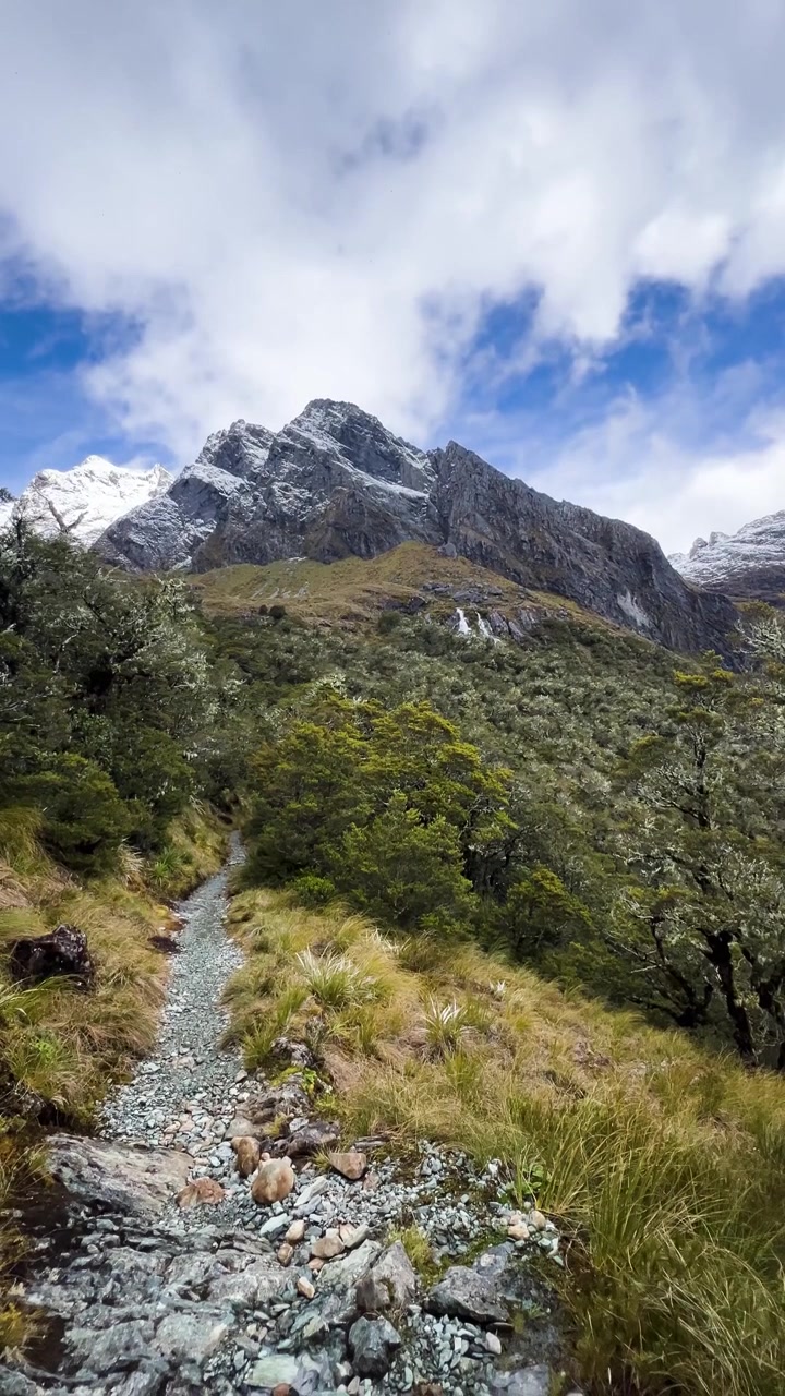 Vertical footage of rocky alpine trail winding toward jagged snow-dusted mountains on Routeburn track.