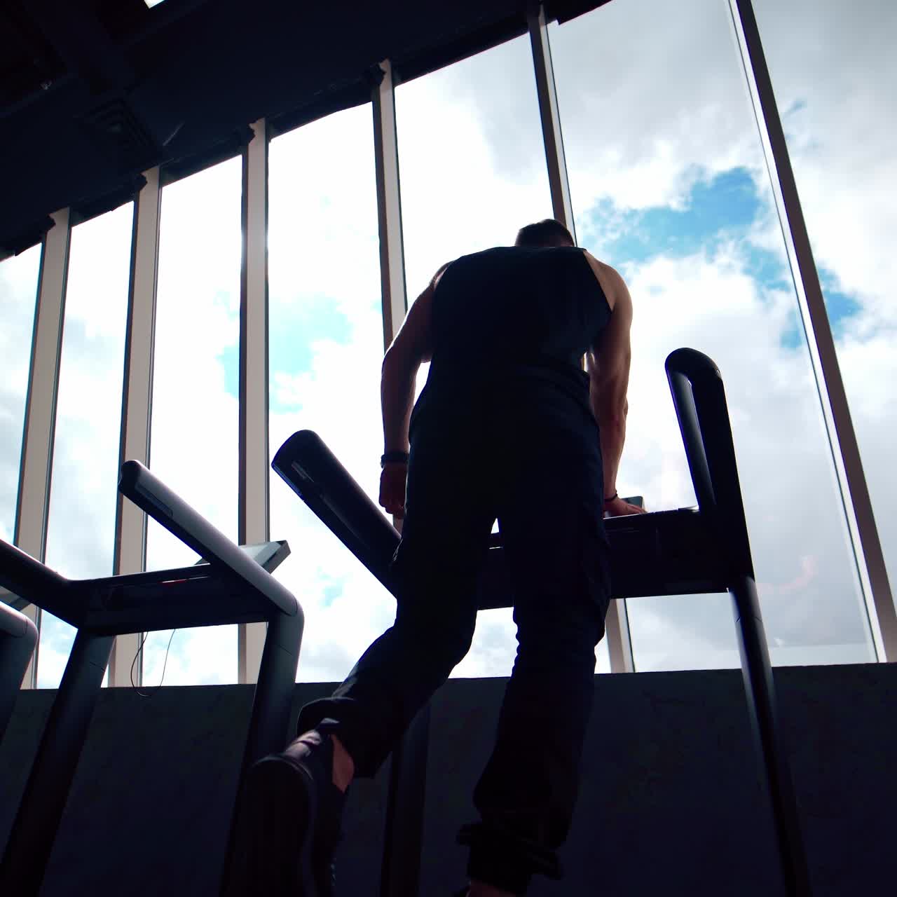 Sportsman on treadmill in gym. Guy in sportswear standing on a treadmill and doing running exercise on window background. Back view from below
