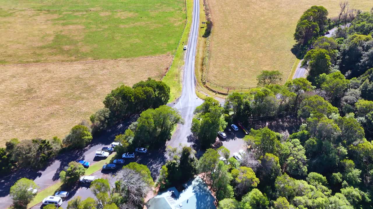 Aerial footage shows a drone smoothly flying above a winding rural road, passing a forested area, open fields, and a large building in bright daylight