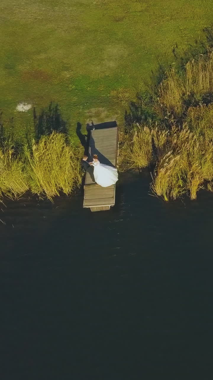 distant newlywed couple stands on small wooden pier near large tranquil river on autumn day aerial view