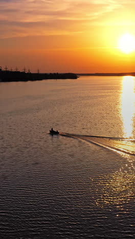 aerial view over the water and motor boat floating at sunset. beautiful view on the setting orange sun that reflects in water. boat on the river moves to the industrial factory. vertical video