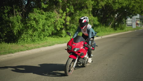 a female motorcyclist navigates her red power bike skillfully while standing and then sits down as she continues her ride. trees are seen lining the road