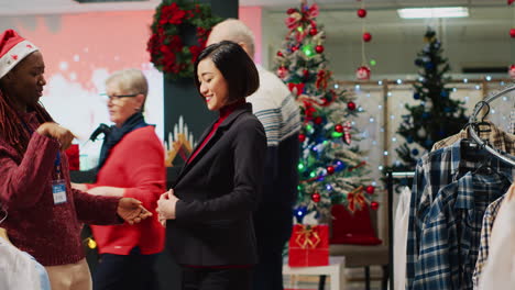 Mujer-De-Negocios-Haciendo-Piruetas-Mientras-Prueba-Una-Chaqueta-Elegante,-Pidiendo-Opinión-Al-Trabajador-Antes-De-Comprarla.-Cliente-Y-Empleado-En-Navidad-Adornan-La-Tienda-De-Ropa-Durante-La-Temporada-De-Vacaciones-De-Invierno