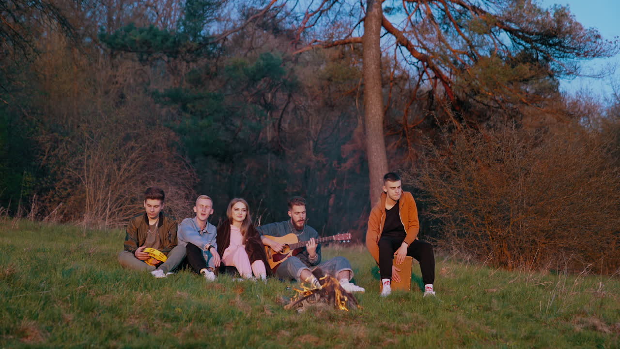 Evening near the fire outdoors. Group of friends sitting near the campfire in the forest. Young friends singing to the guitar in nature.