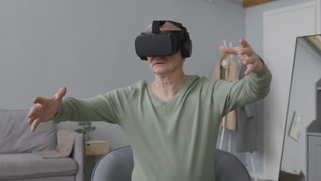 hombre mayor que usa gafas de realidad virtual y mueve las manos mientras se sienta en una silla en una sala de estar moderna 1