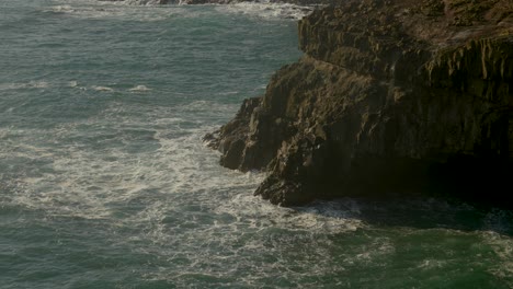 Aerial-view-of-ocean-waves-crashing-against-rocky-cliff-coast-during-sunny-day-on-Faroe-Island