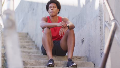 african american man exercising in city, sitting on steps and tying shoelaces