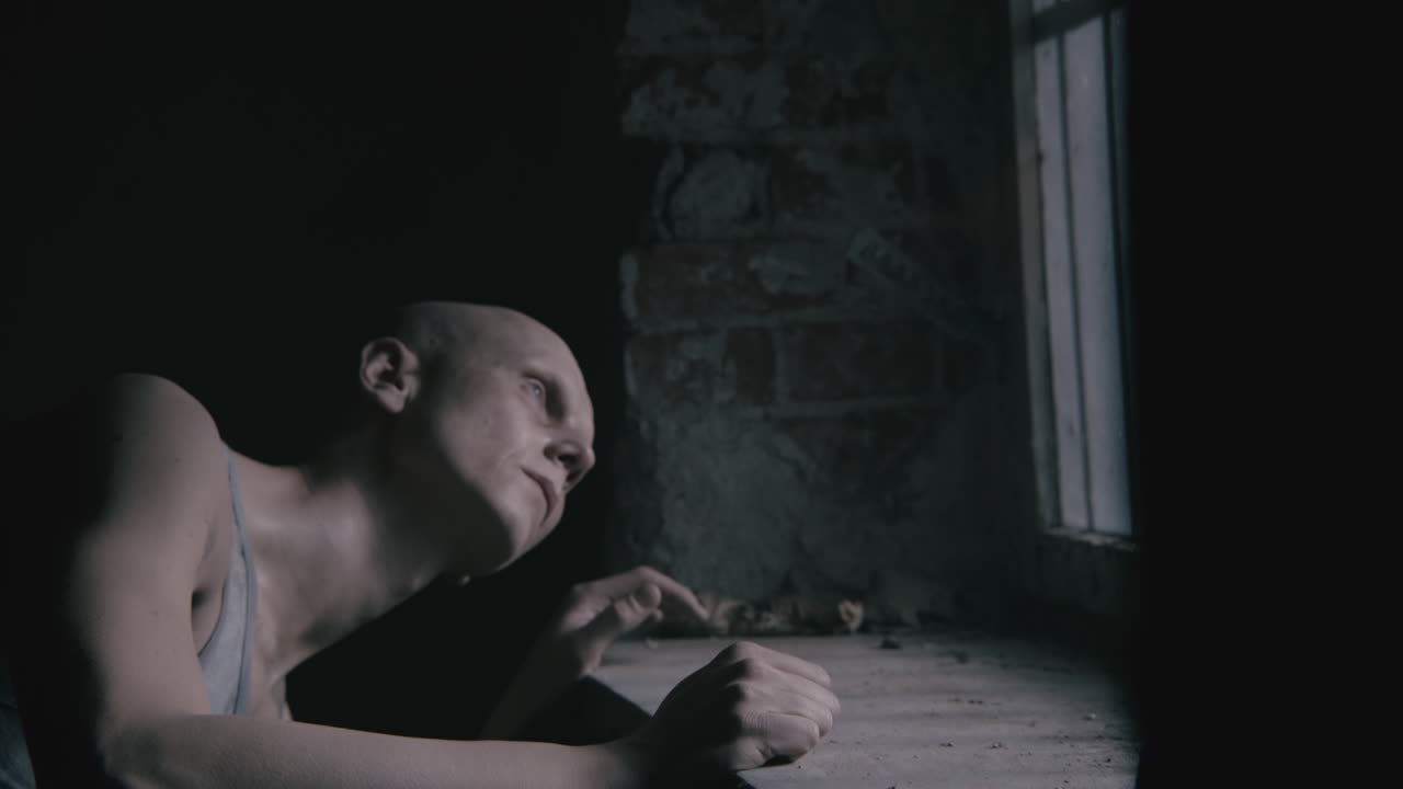 Pale bald skinny man looks through the window in the creepy old bathroom at night