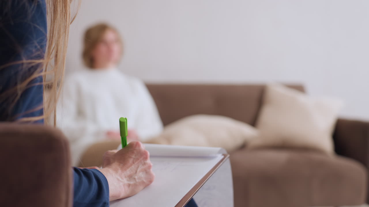 Patient seated on sofa communicates during session while attentive counselor listens and writes on notepad, illustrating emotional dialogue, therapeutic support, and calm professional interaction
