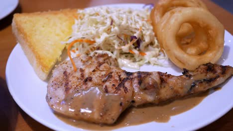 close-up of a platter of chicken steak in barbecue sauce, served alongside some onion rings, a coleslaw salad, and a slice of buttered toasted bread