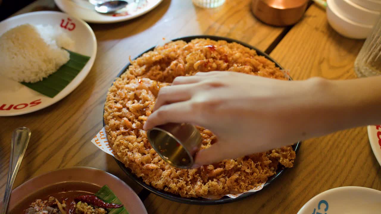 Hand pours chili sauce over golden fried shrimp pancake in bright, inviting Thai restaurant setting