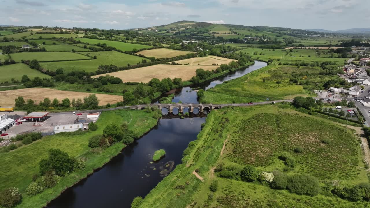 Clady Bridge, County Tyrone, Northern Ireland, June 2023. Drone panoramic orbit ascend clockwise showcasing arched bridge crossing River Finn with clouds reflecting in water and both villages visible.